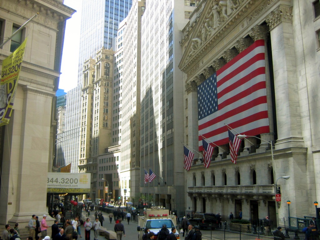Transcendent Prosperity - The New York Stock Exchange building with a large American flag displayed on its facade, surrounded by skyscrapers and people on the street.
