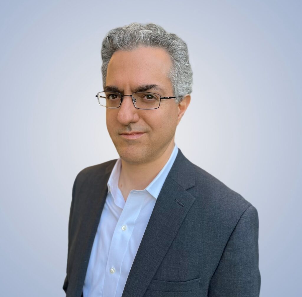 Transcendent Prosperity - A man with gray hair and glasses wears a dark suit jacket over a white shirt, standing against a plain light background.
