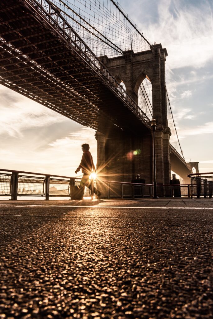 Transcendent Prosperity - A person walks a dog under the Brooklyn Bridge at sunrise, with sunlight streaming through the bridge's arch and the pavement in the foreground, capturing a moment of transcendent prosperity.